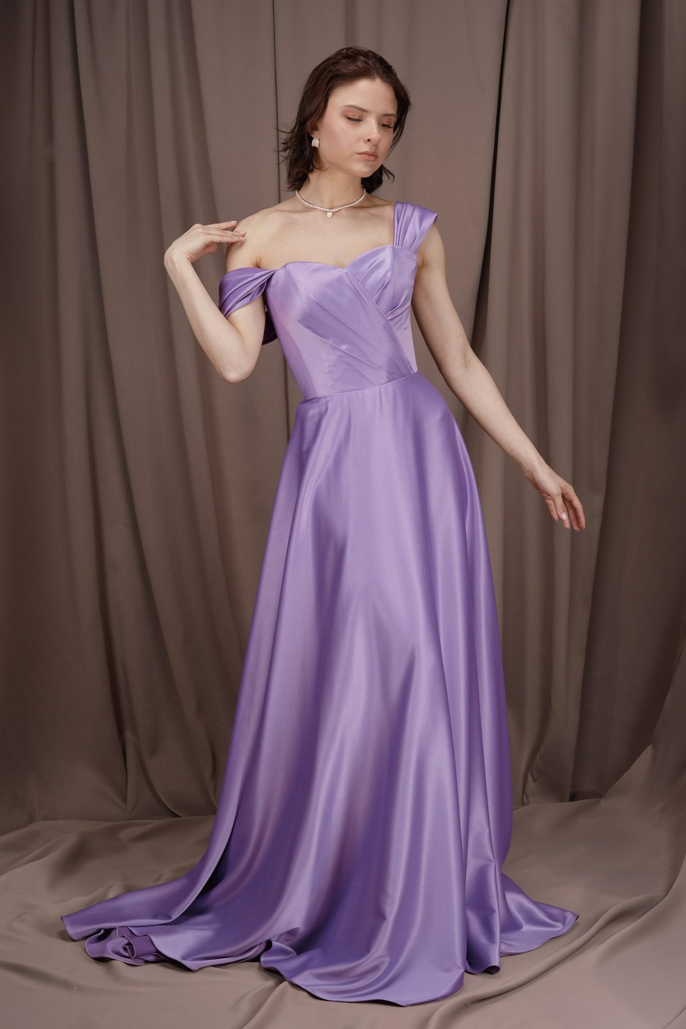 Девушка в длинном вечернем платье сиреневого цвета. У платья открытые плечи и лиф с драпировками. Юбка платья а-образная и ниспадает красивыми складками до пола.