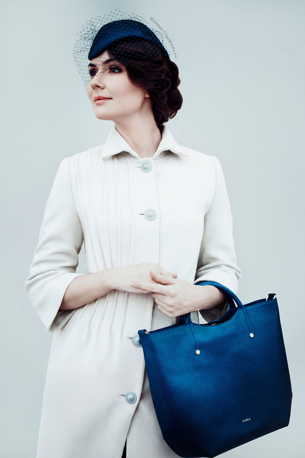 Синяя шляпка с вуалью и прической на девушке в светлом пальто с синей сумкой