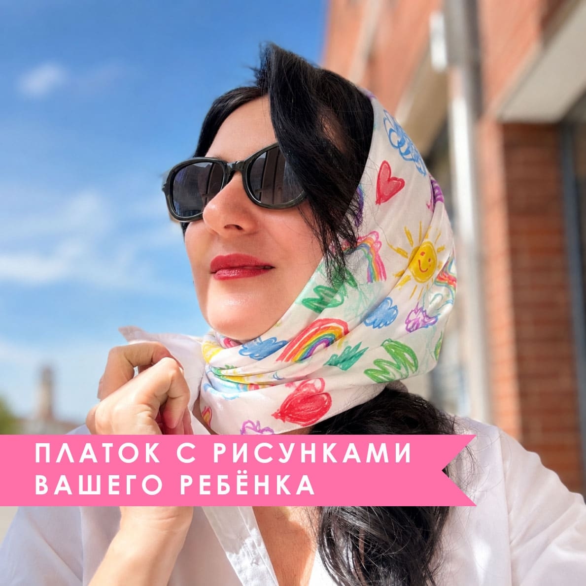 Брюнетка в солнцезащитных очках позирует на улице в светлом шёлковом платке на заказ, украшенном яркими детскими рисунками радуги, солнца и сердец, с розовой рекламной плашкой «Платок с рисунками вашего ребёнка».