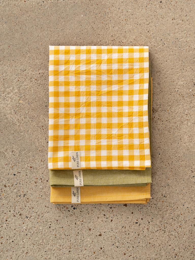 Набор кухонных полотенец в тёплых оттенках: горчичная клетка, оливковый и жёлтый цвета, аккуратно сложены на каменной столешнице.
